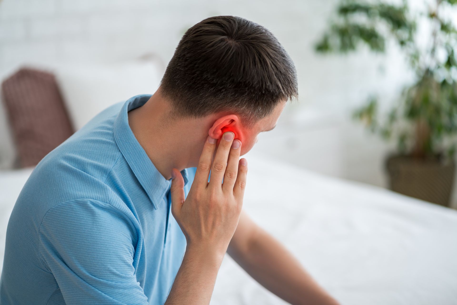 Man in blue shirt touches his red ear, appearing to be in pain, indoors.