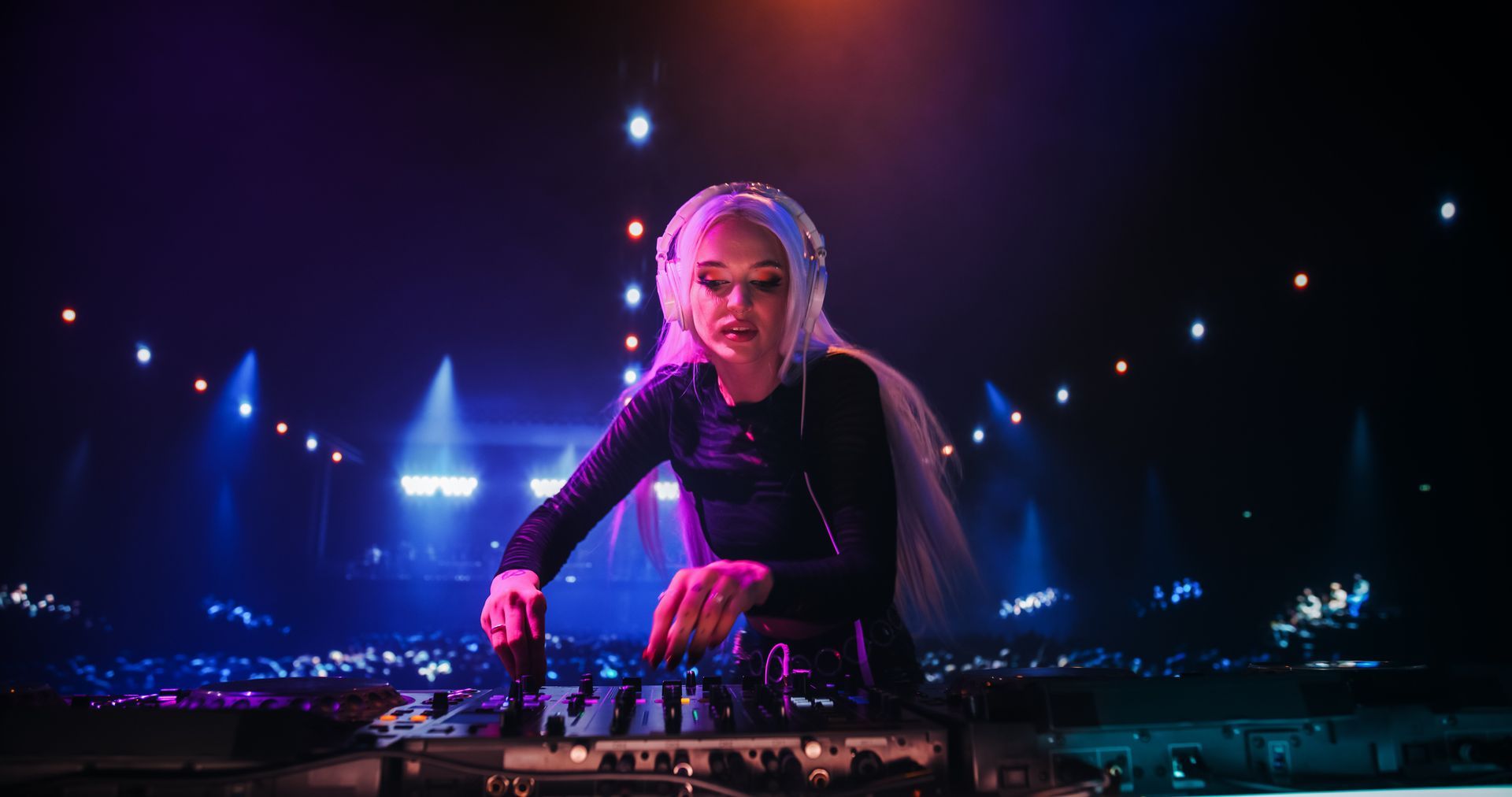 Woman DJ at a concert, mixing music with a dark outfit and headphones, surrounded by lights and a crowd.