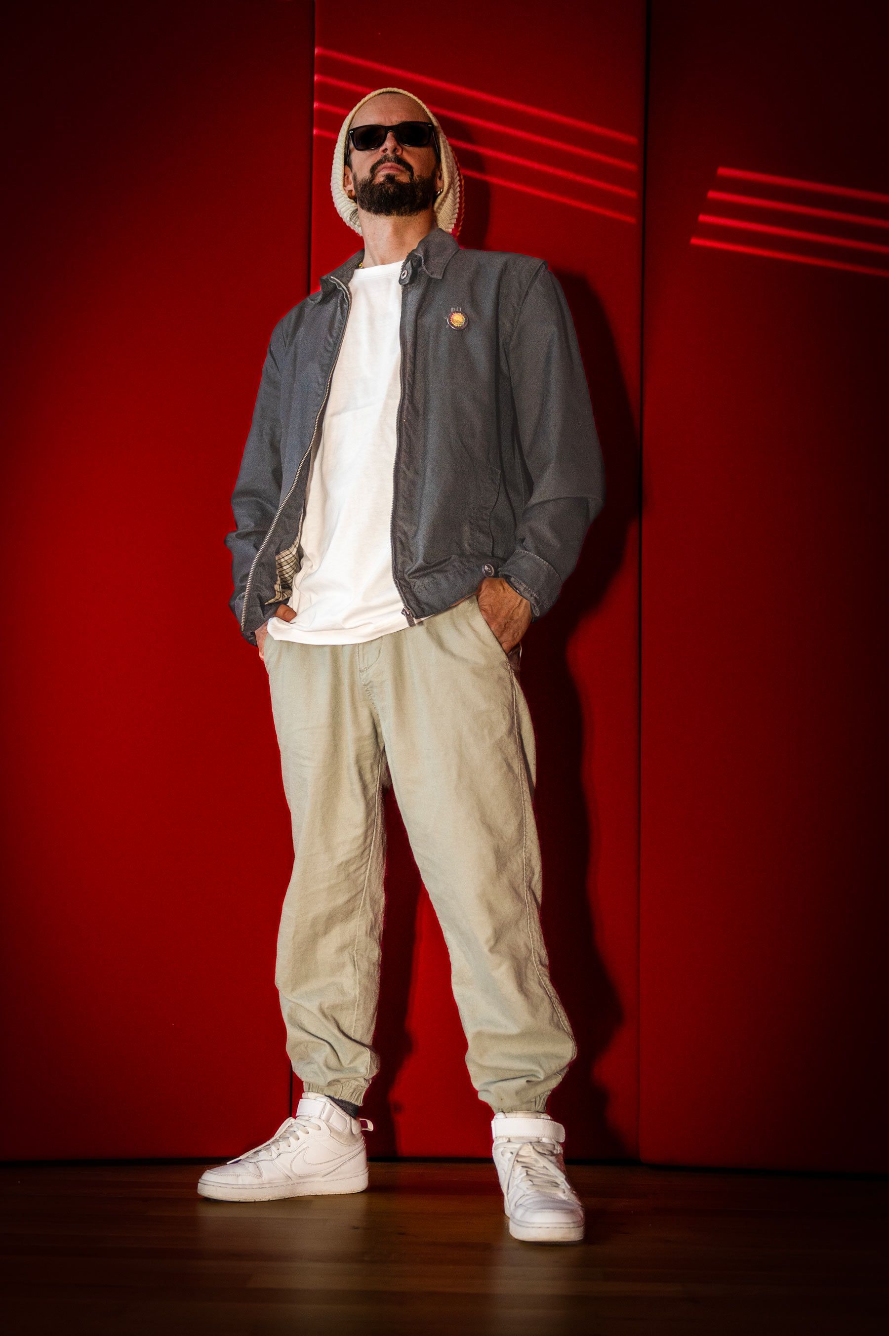 Un uomo con la barba e gli occhiali da sole è in piedi davanti a un muro rosso.