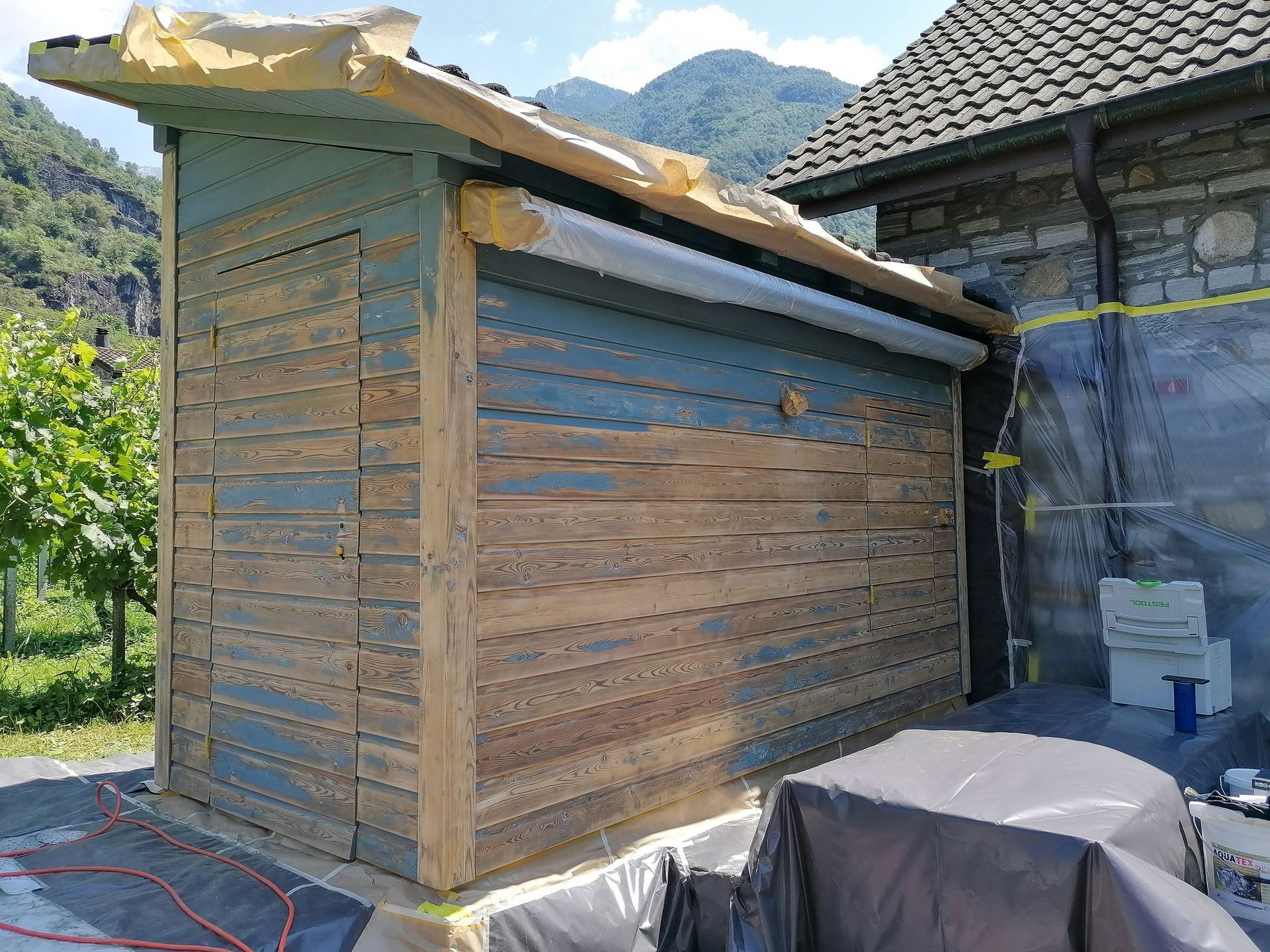 Davanti ad un edificio in pietra si sta ridipingendo una tettoia di legno.