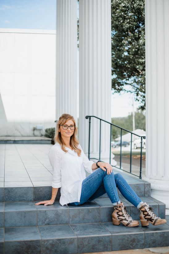 Rachel  is posing for a headshot, sitting on stone steps in front of white pillars smiling.