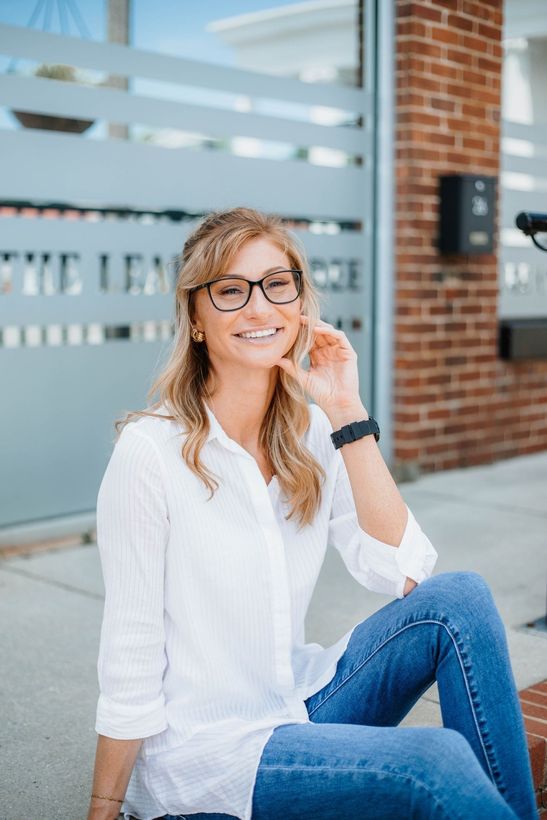 A woman is posing for a headshot, sitting on the sidewalk smiling.