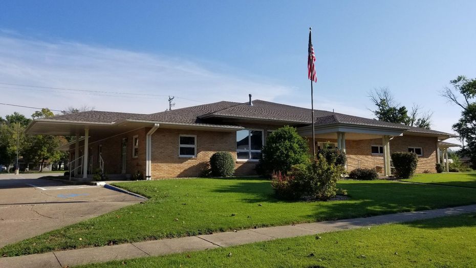 Low brick building with awning, green lawn, American flag on pole.