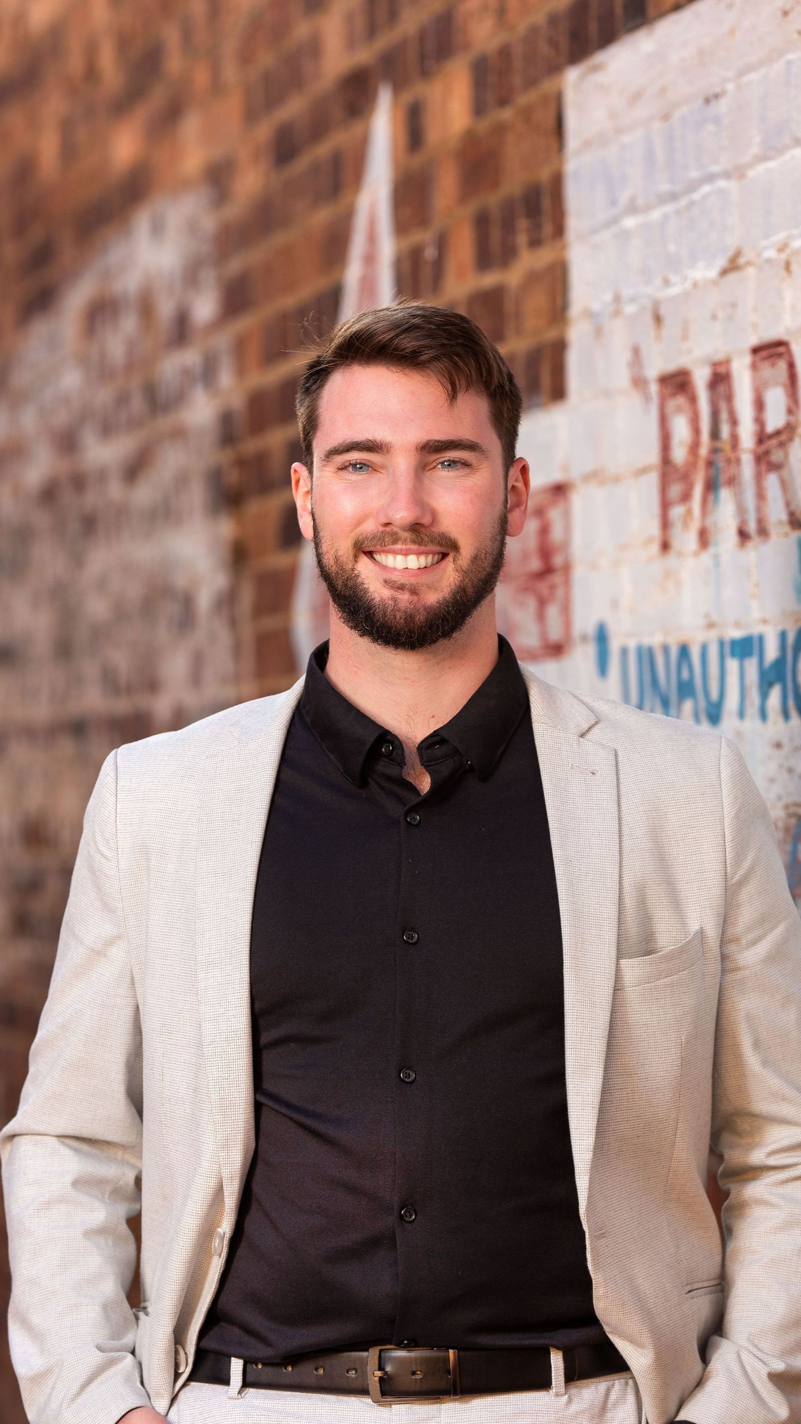 Jai Warner n a suit and black shirt is standing in front of a brick wall in a business portrait.