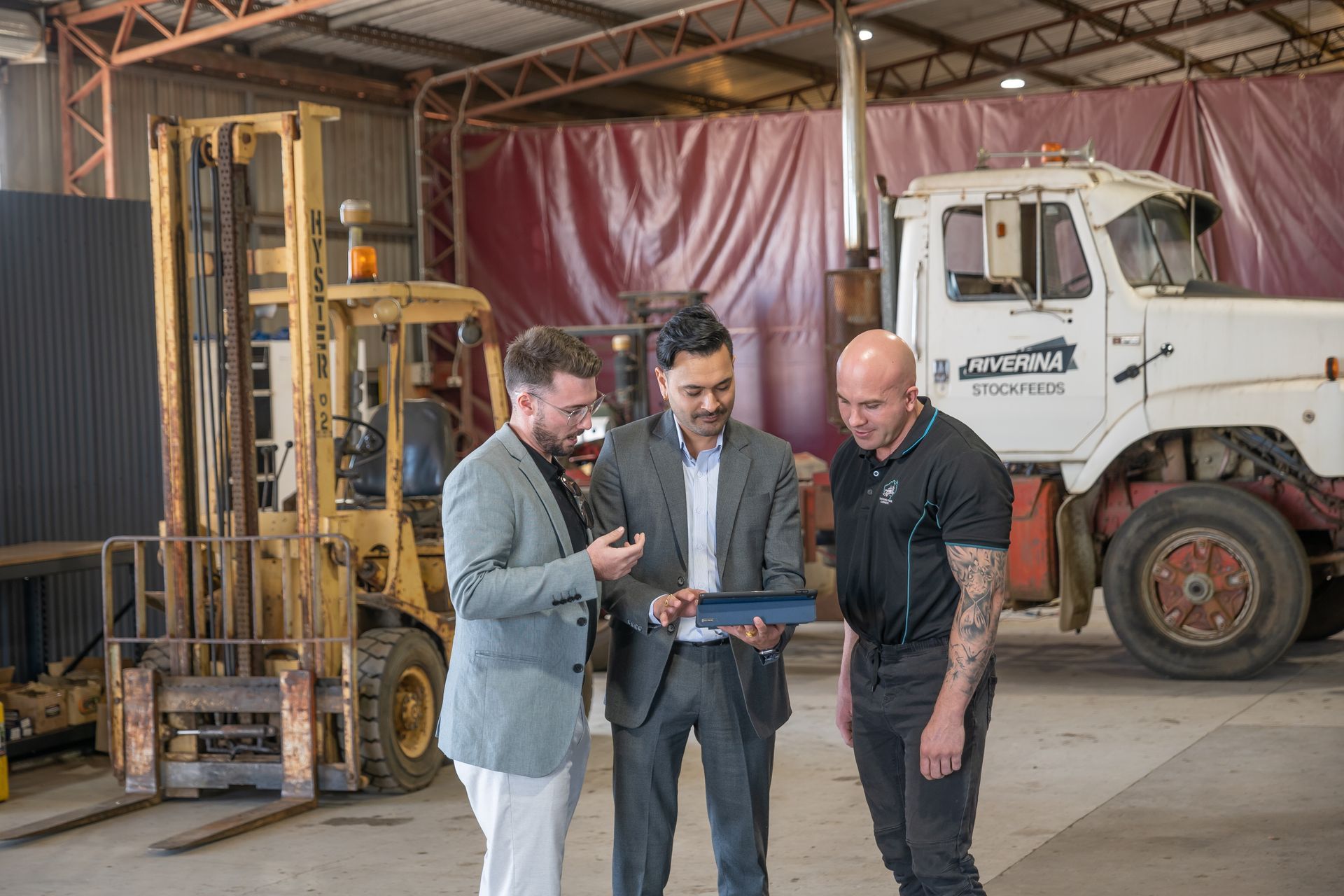 three men are standing in front of a forklift and a truck in a warehouse reviewing digital marketing reports.