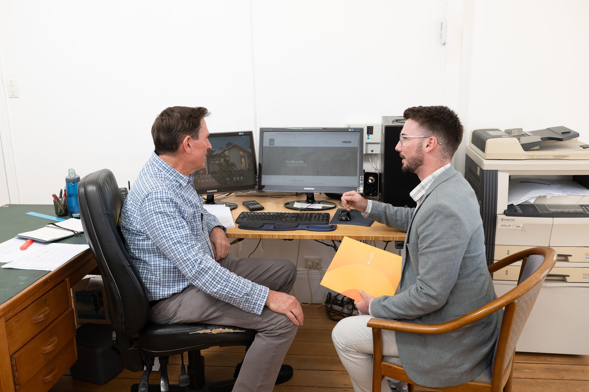 a man sitting at a desk talking to another man in front of a computer