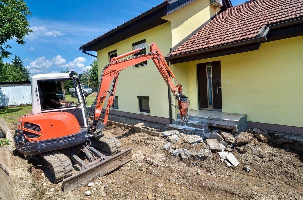 A Small Excavator Is Sitting In Front Of A Yellow House — Glen Tier Bobcat & Truck Hire In Innisfail, QLD