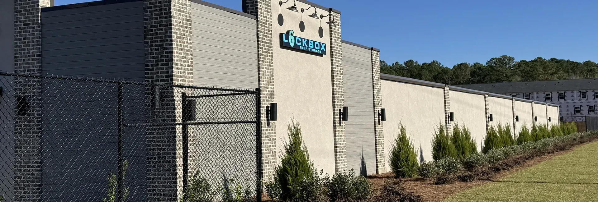A light-colored building with a logo, featuring a row of green bushes, with a clear blue sky background.