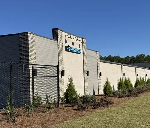 A light-colored building with a logo, featuring a row of green bushes, with a clear blue sky background.