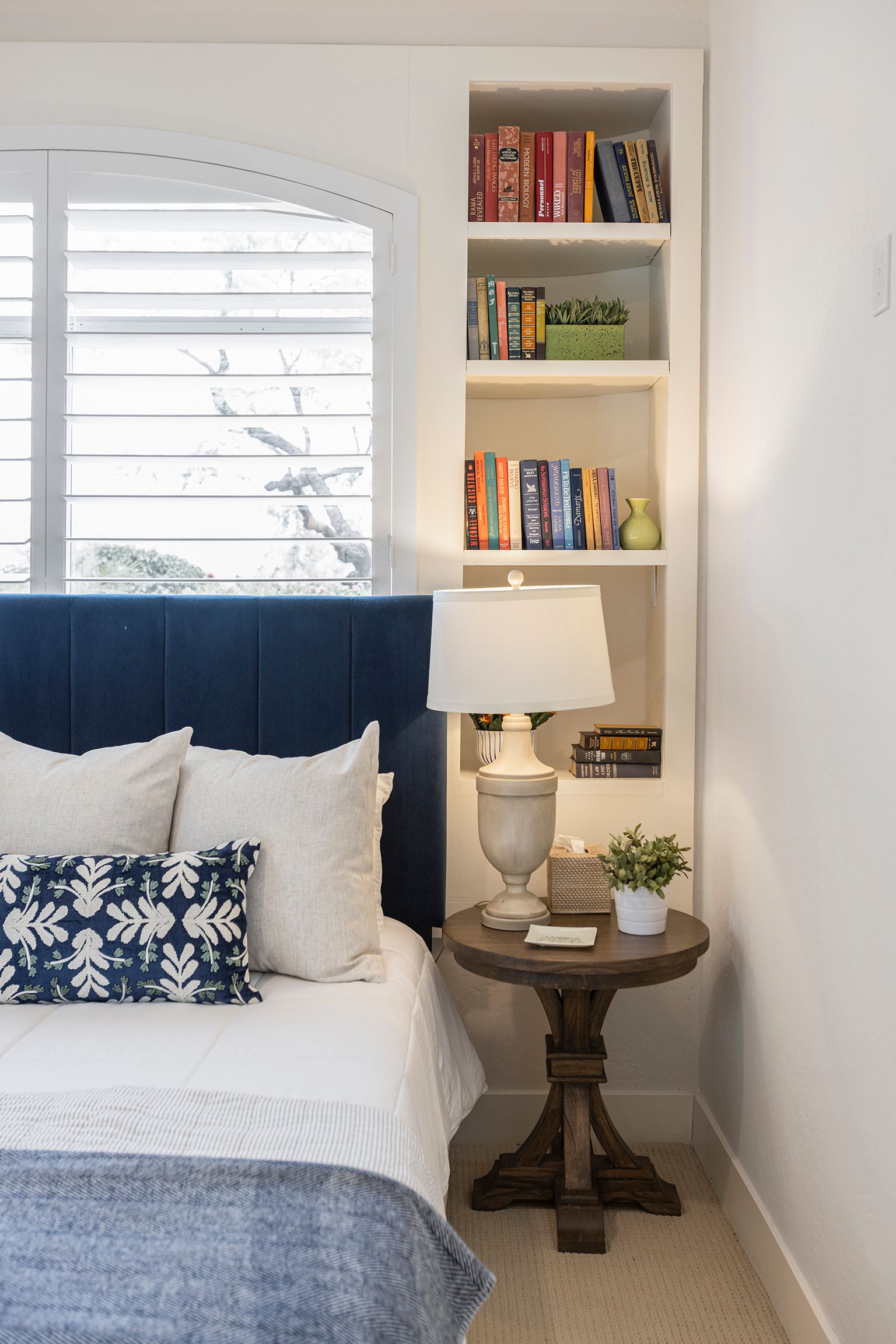 A bedroom with a bed , nightstand , lamp and bookshelves.