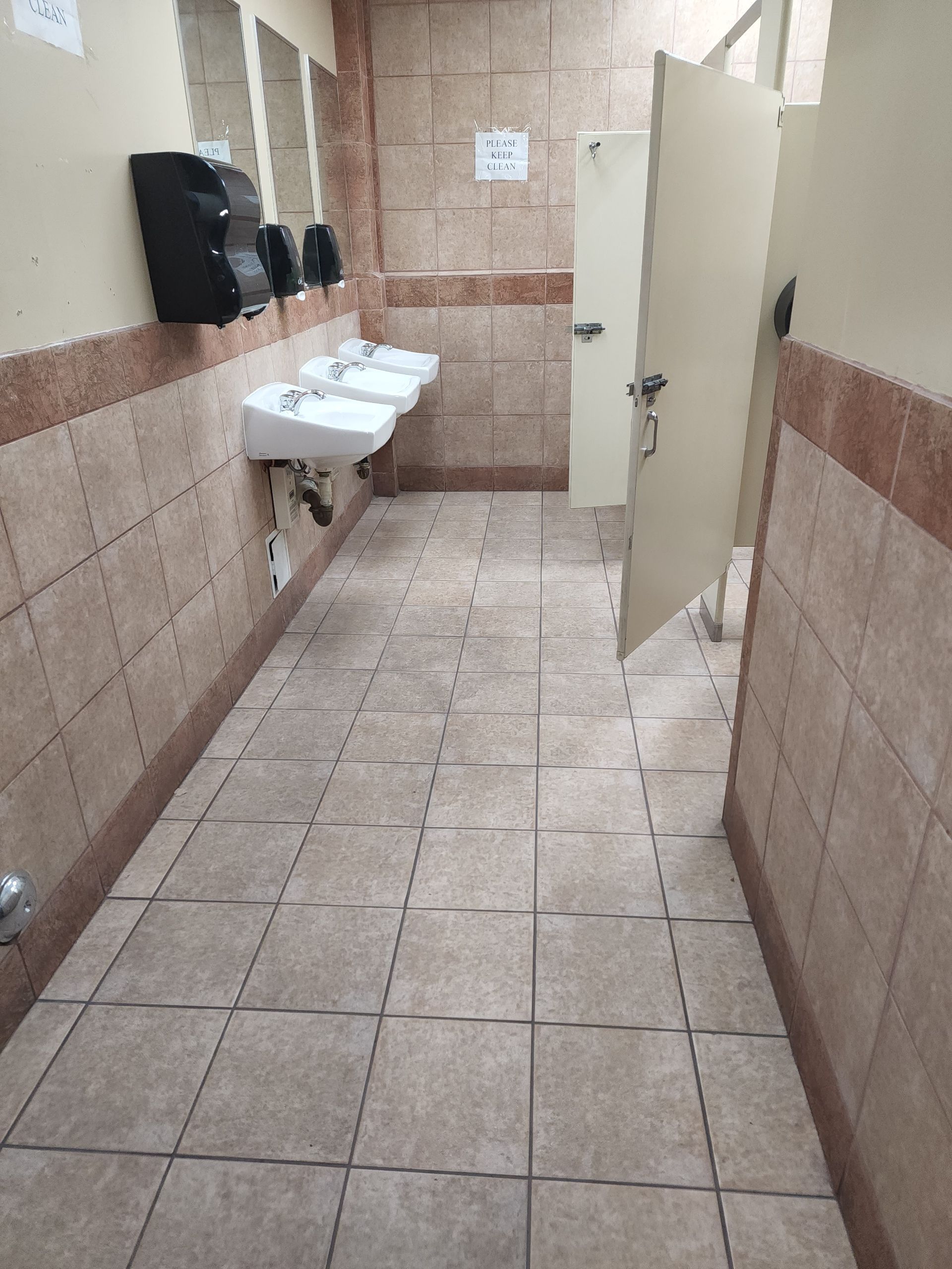 Public restroom with tan tile floor and walls, white sinks, and open stall door.