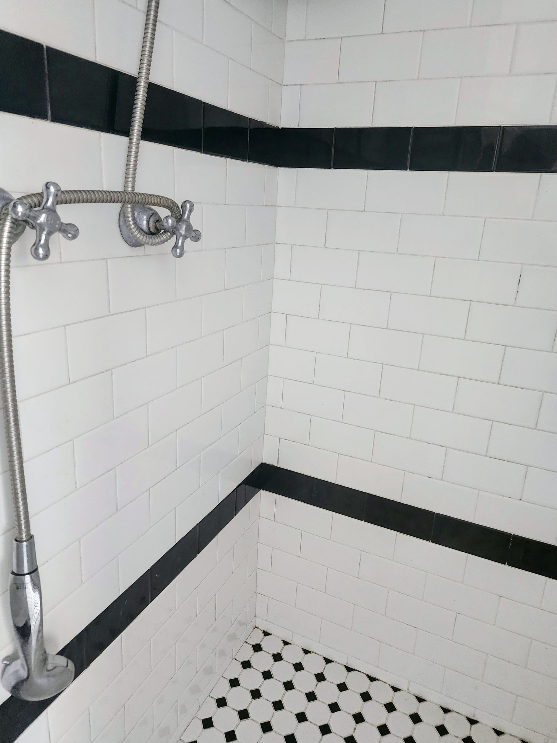 White-tiled shower stall with chrome fixtures, black trim, and black-and-white mosaic floor.
