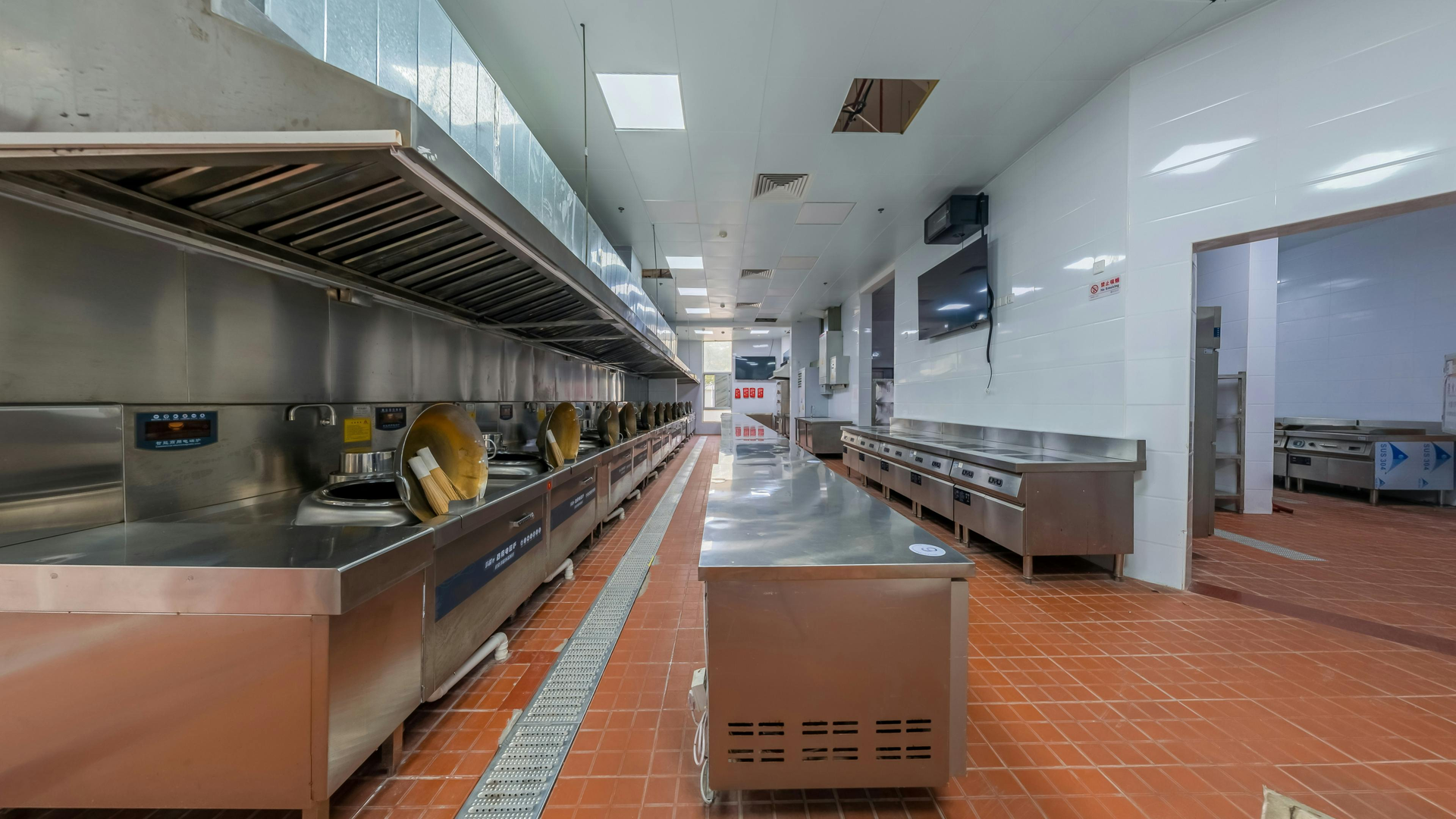 Long commercial kitchen with stainless steel counters and appliances, tile floor, and overhead ventilation.