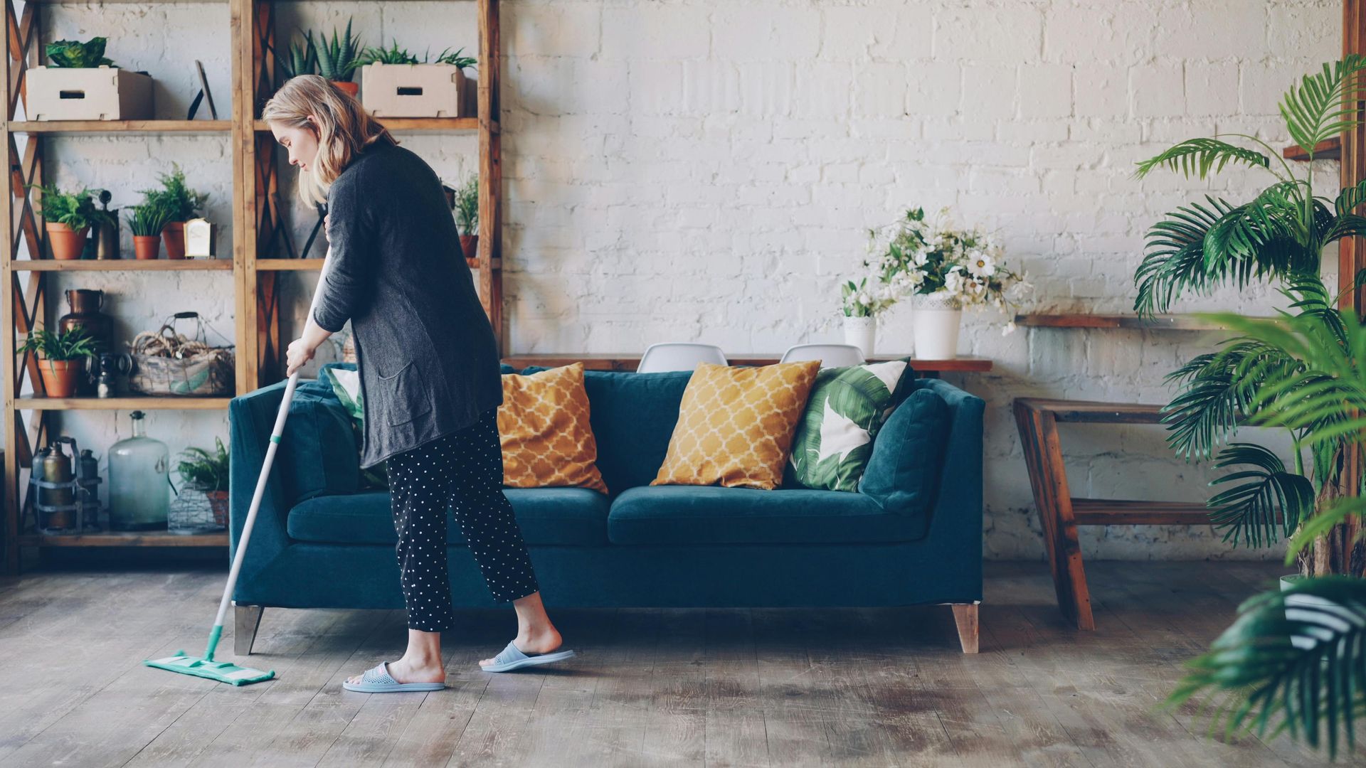Person mopping a cozy living room with teal sofa, shelves, and plants.