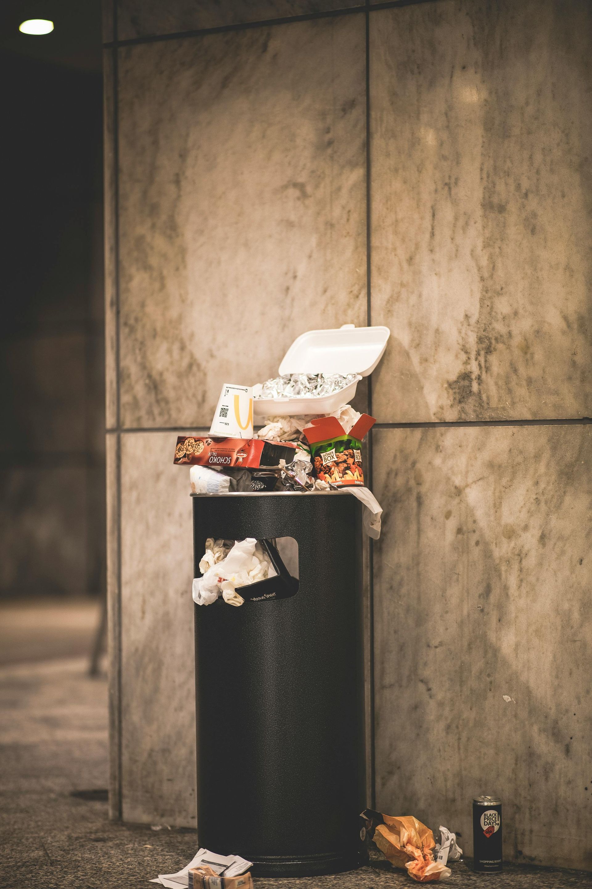 Overflowing black trash can against a concrete wall, with scattered trash and a can on the ground.