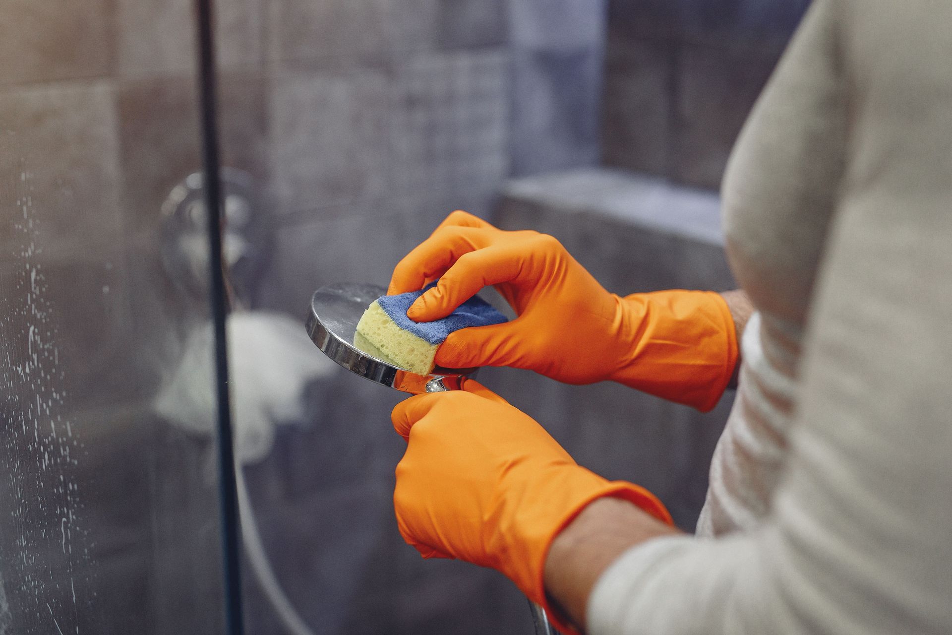 Person in orange gloves cleaning a glass surface with a sponge in a shower area