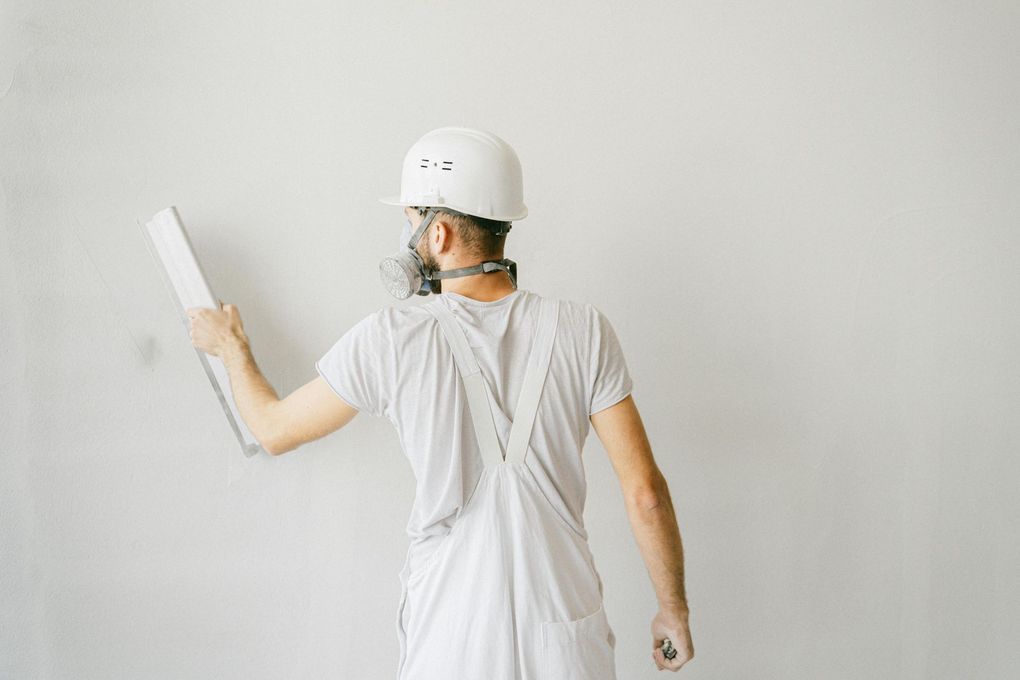 Person in white overalls and hard hat smoothing wall with a trowel.
