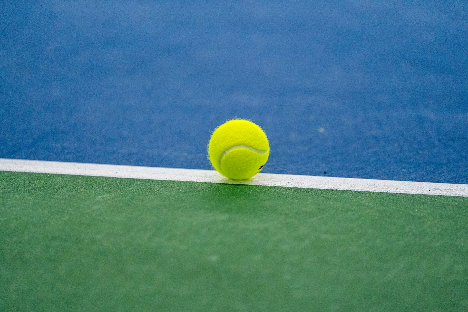 A tennis ball is sitting on the line of a tennis court.