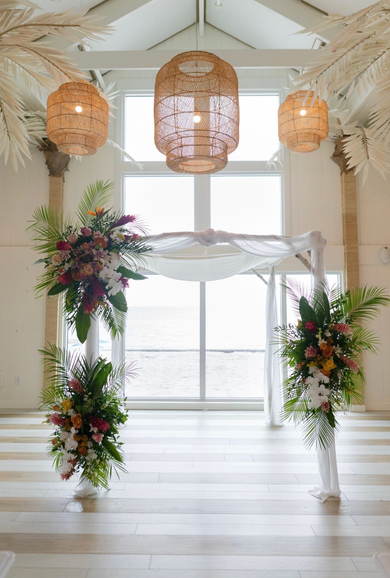 Wedding ceremony space with floral arch, large window, and woven pendant lights.