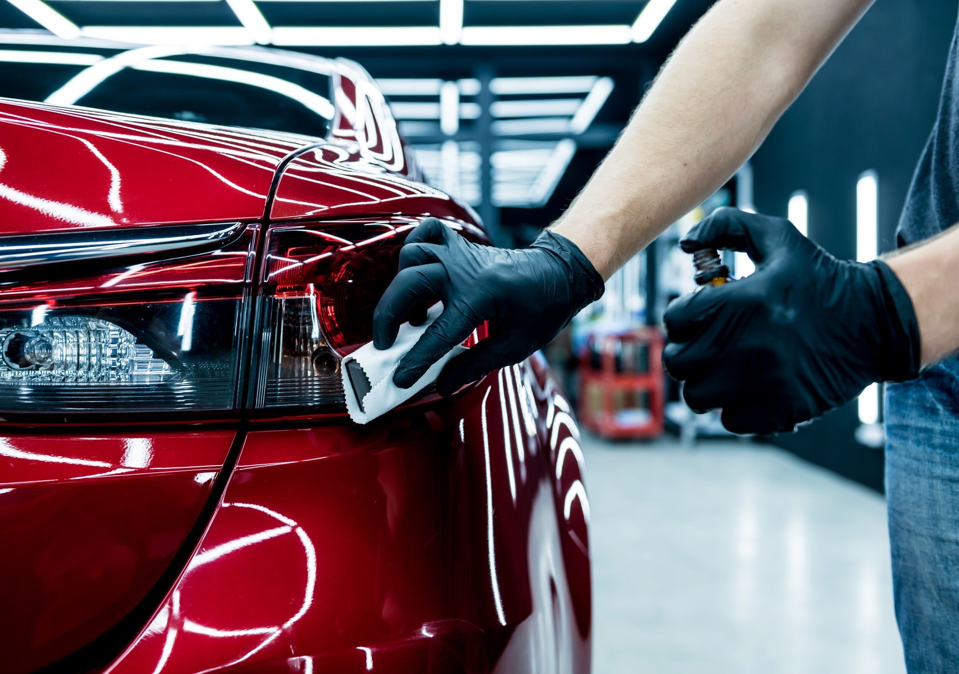 A man wearing black gloves is polishing a red car in a garage.