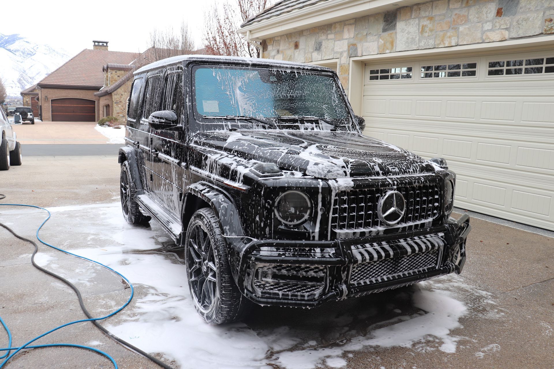 A black mercedes benz g63 is covered in foam in a driveway.