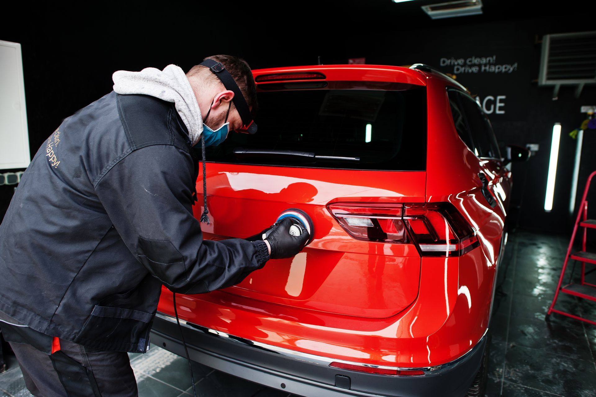 A man wearing a mask is polishing a red car in a garage.