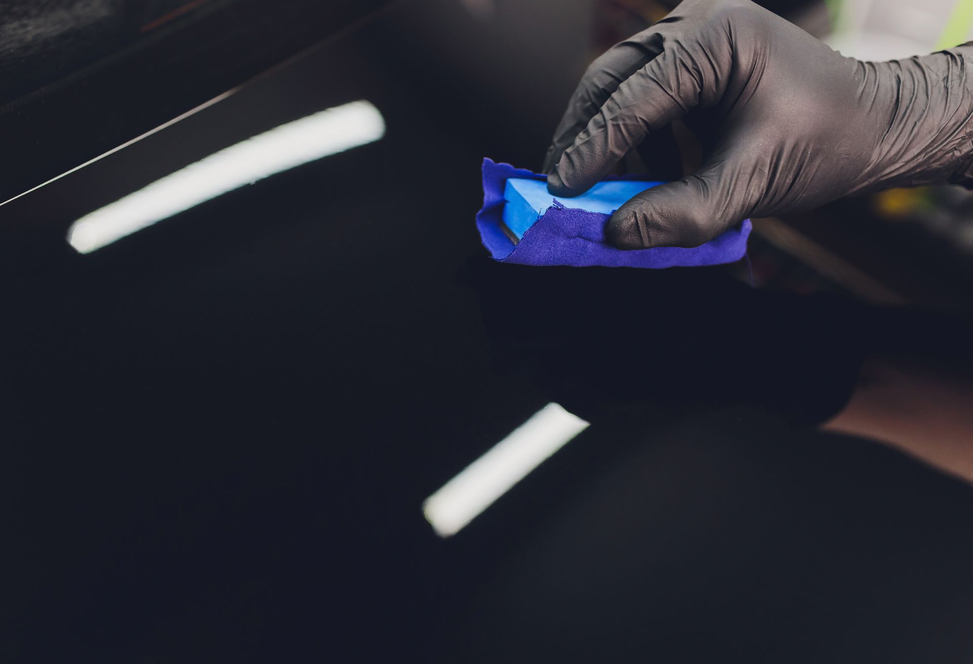 A person wearing black gloves is polishing a car with a blue sponge.