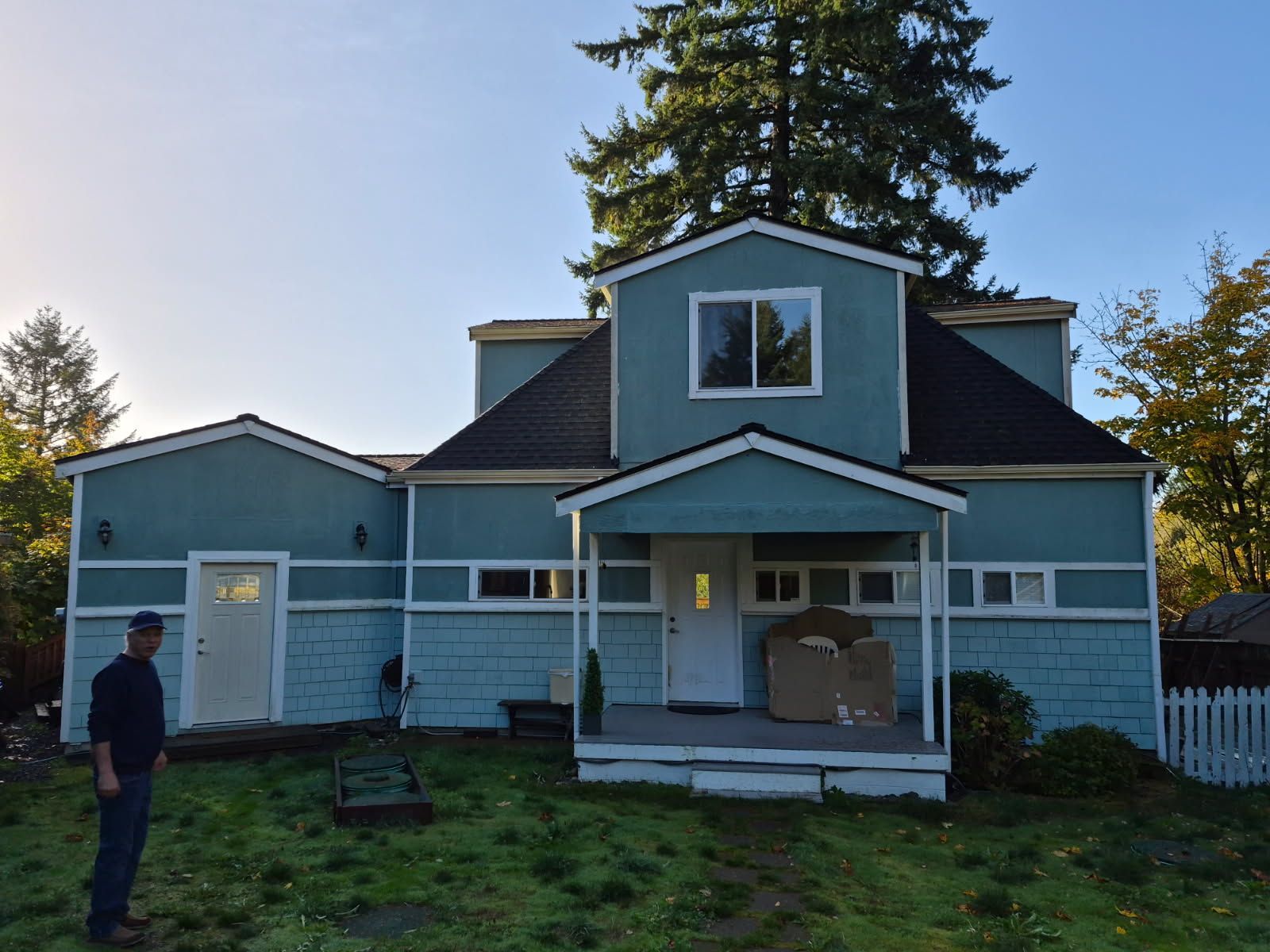 Man stands in front of a turquoise house with a white porch and white trim. Overcast, grassy yard.