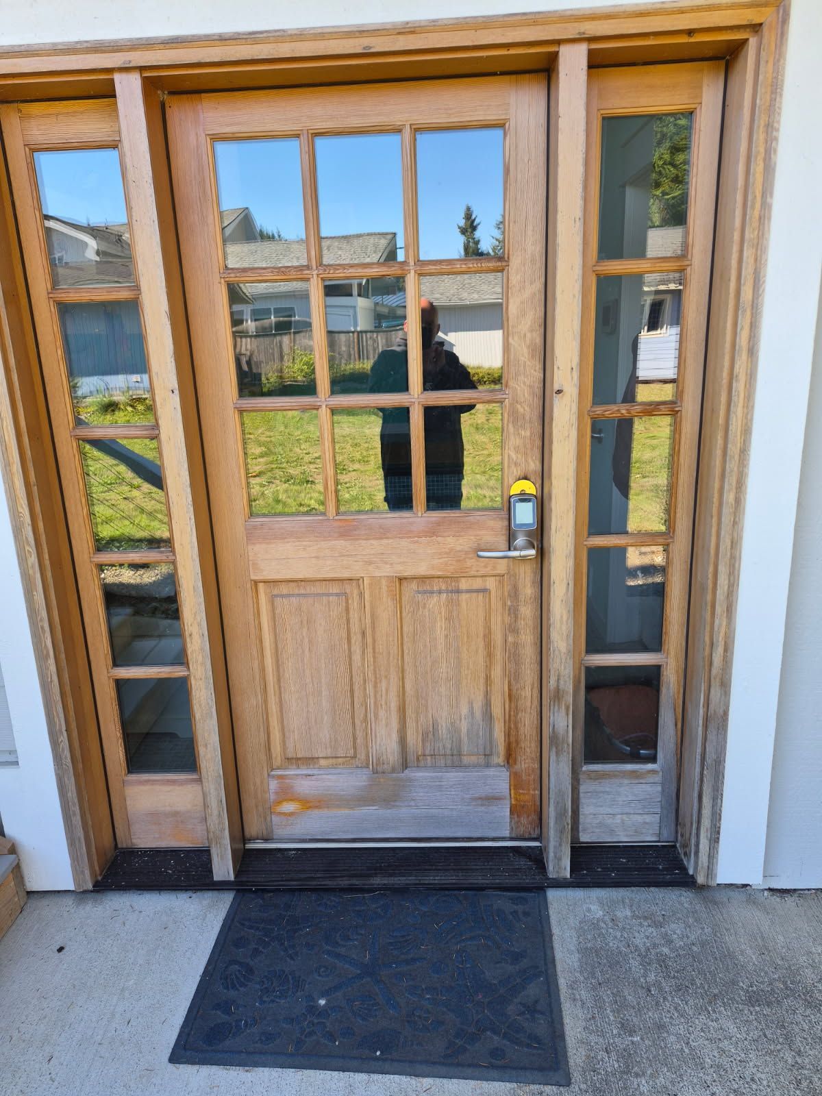 Wooden front door with glass panes and side windows; a person's reflection is visible.