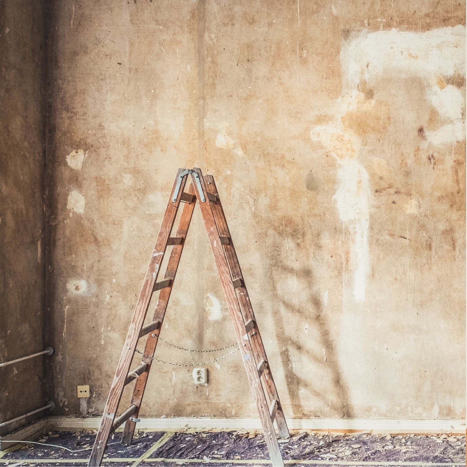 A wooden stepladder stands in the center of an empty room with unfinished walls and a dark, debris-covered floor.