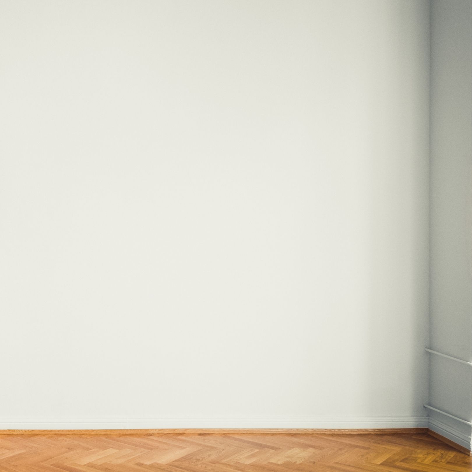 A plain white interior wall meeting a corner above a light-colored wooden floor with a herringbone pattern.