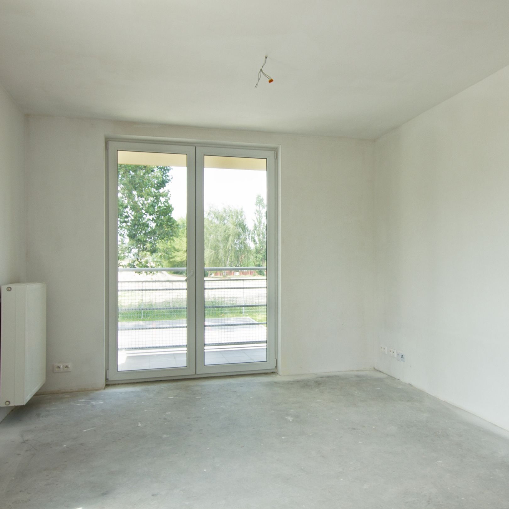 An empty, unfinished room with white walls, a gray concrete floor, a radiator, and a glass door leading to a balcony.