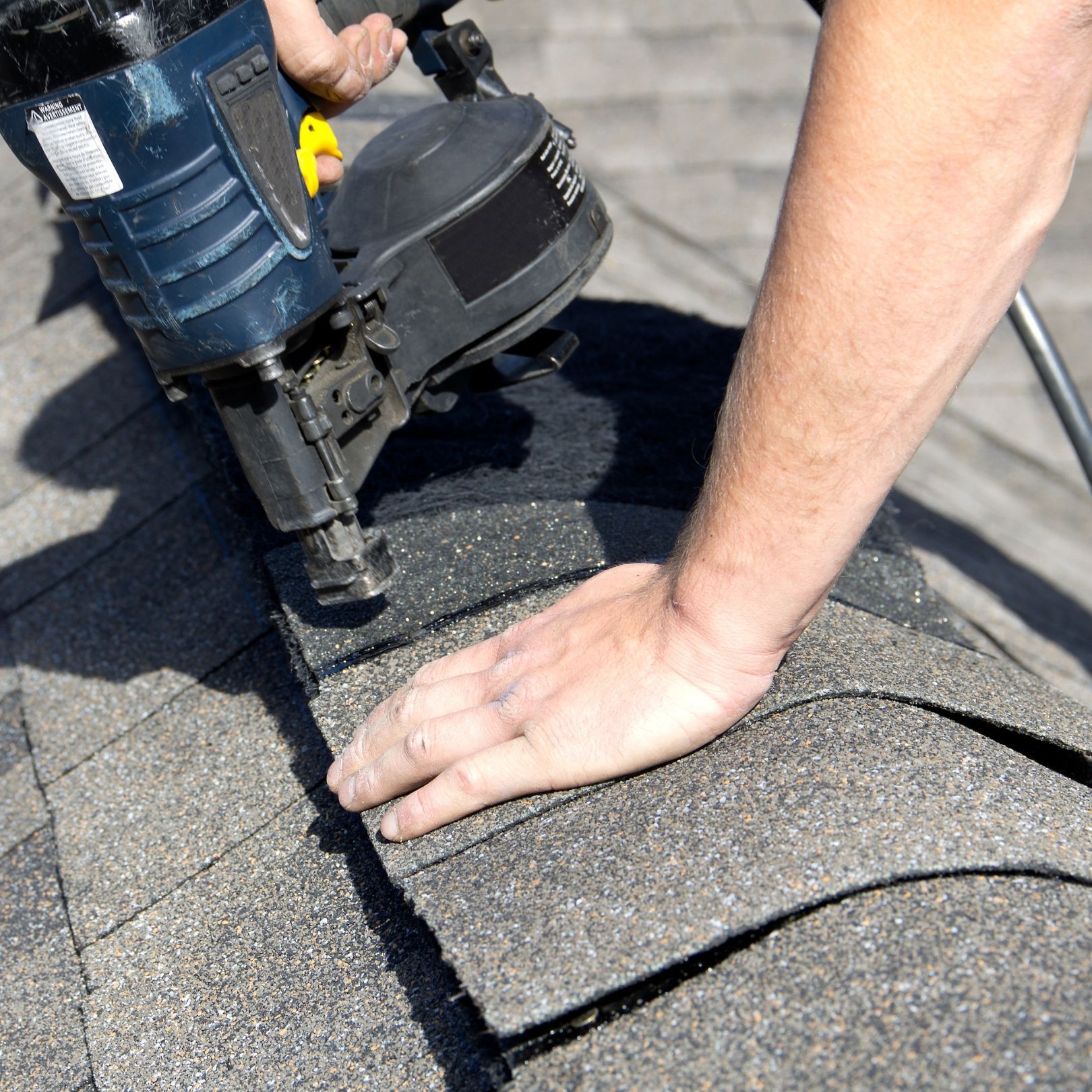 A person uses a pneumatic nail gun to secure gray asphalt shingles onto a sloped roof.