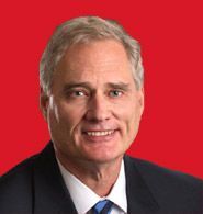 Professional headshot of a man wearing a dark suit, white shirt, and blue tie against a solid red background.