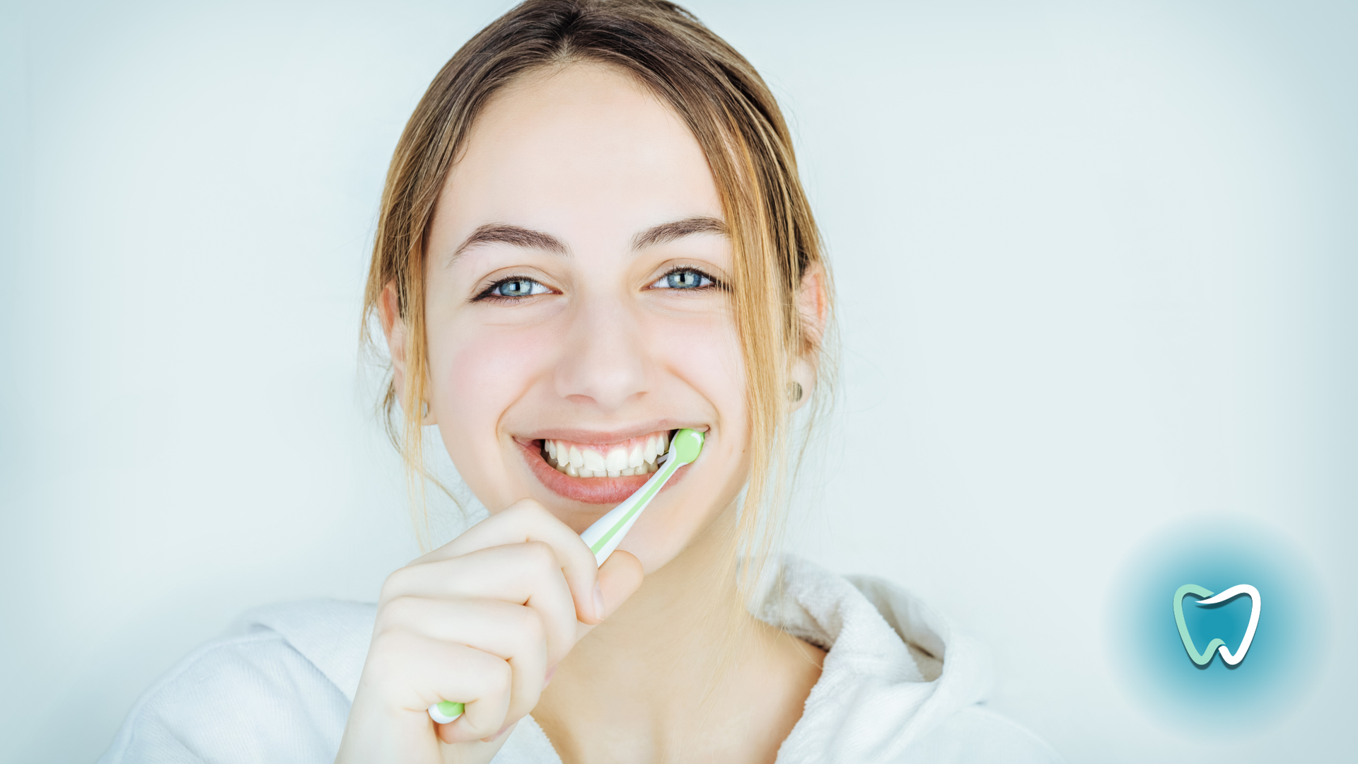 Woman brushing teeth, smiling broadly, light green toothbrush, against a light blue backdrop, dental icon in corner.