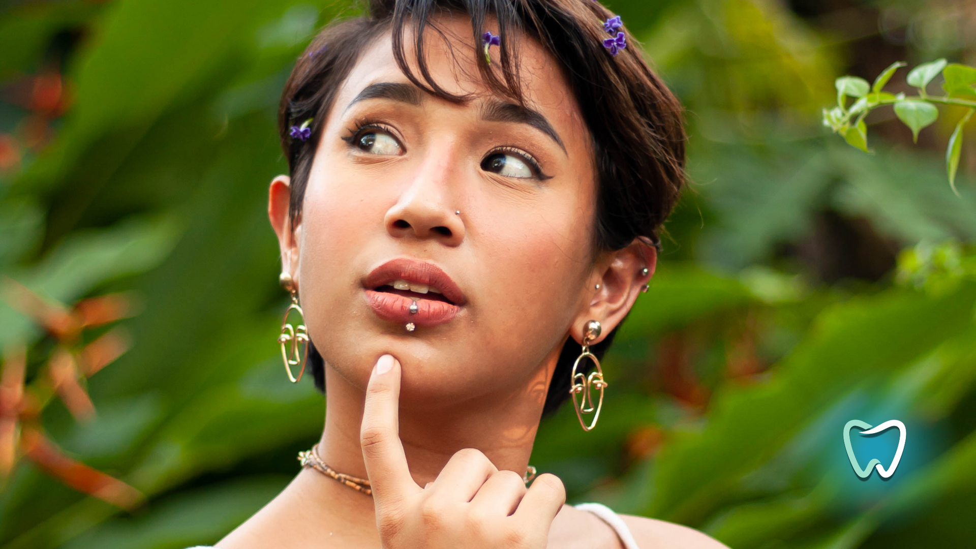 A woman wearing earrings and a necklace is holding her finger to her chin.