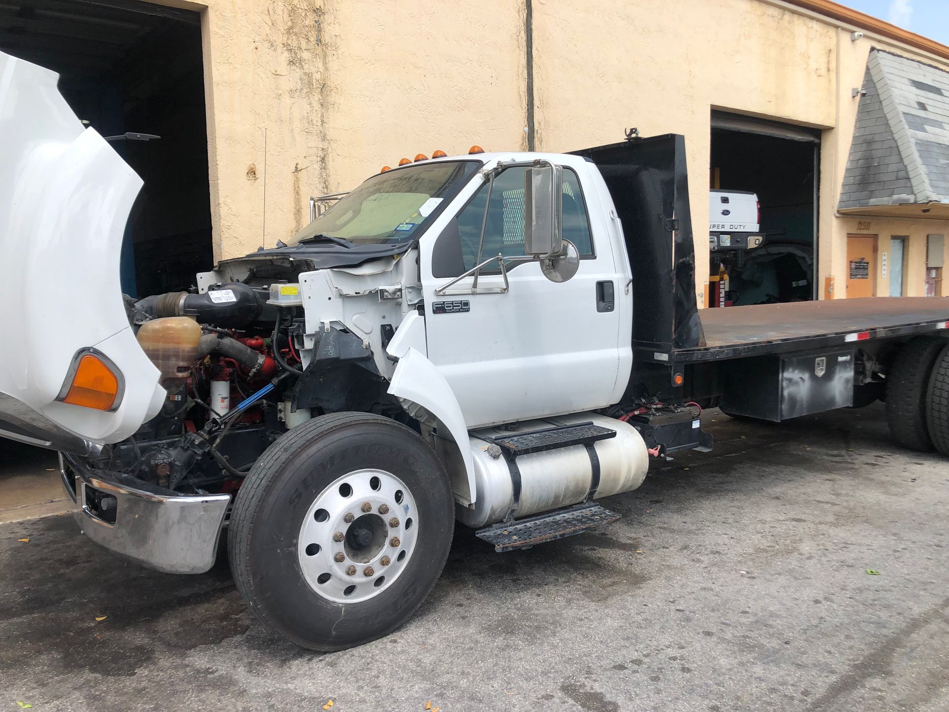 A white flatbed truck with the hood up is parked in front of a building.