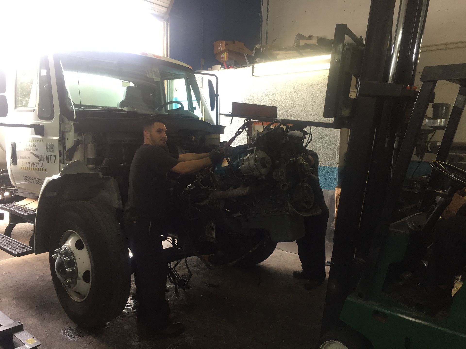 A man is working on a truck in a garage next to a forklift.