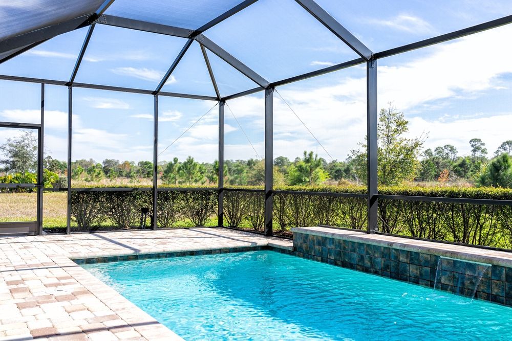 Screened-in pool with blue water and brick patio, framed by black metal and greenery. Sky visible through the roof.