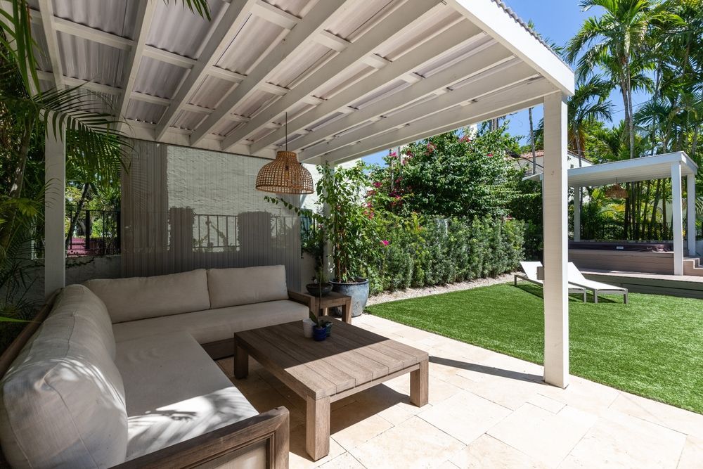Lounge seating under a white pergola in a backyard with green grass, hedges, and a hot tub.