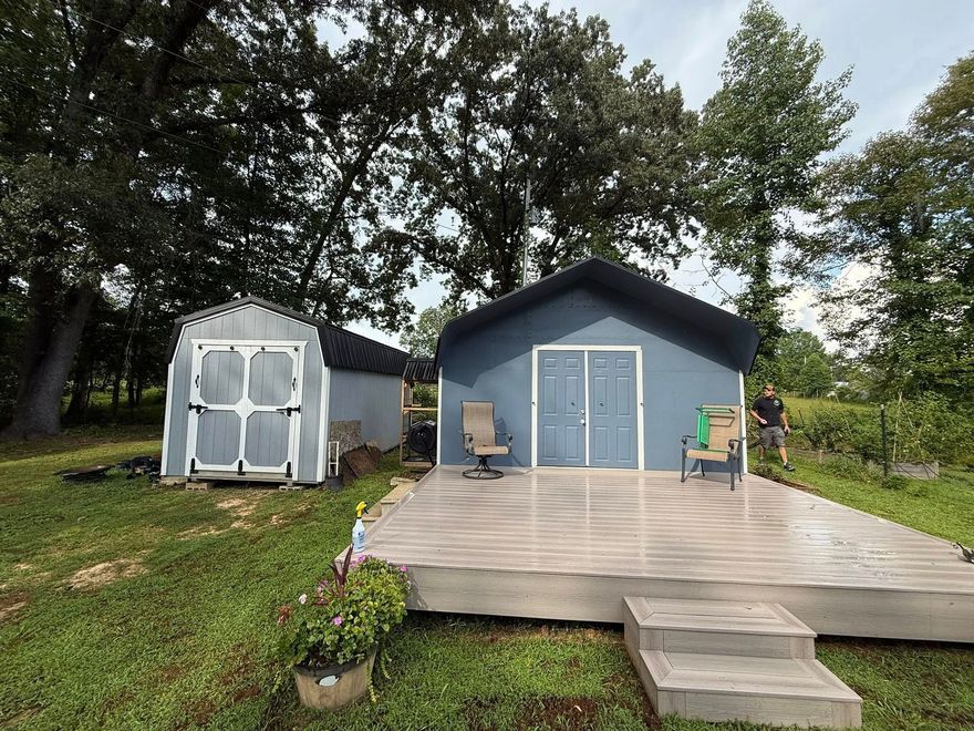 Two sheds in a grassy, wooded yard; one is gray with a large deck and two chairs, the other is white with double doors.