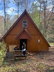 Two people stand at the entrance of a rustic, triangular A-frame cabin in a wooded, leaf-covered area.
