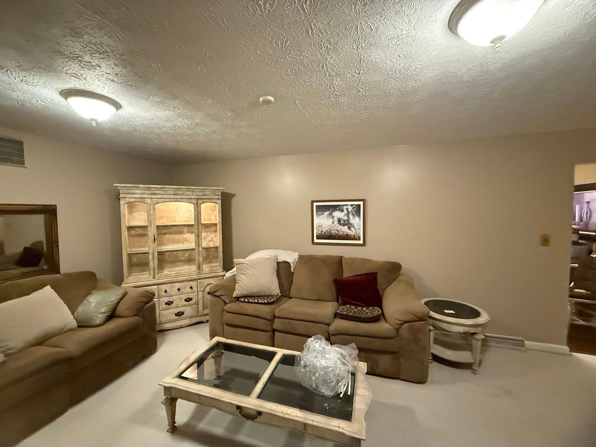 A living room features a tan sofa, glass coffee table, white hutch, and textured ceiling with two flush-mount lights.
