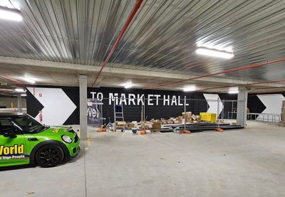 A green car is parked in a parking garage next to a sign that says to market hall — Signworld In Mitchell, ACT