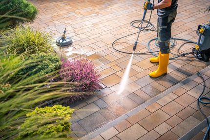 Person in yellow boots pressure washing a brick patio near bushes.