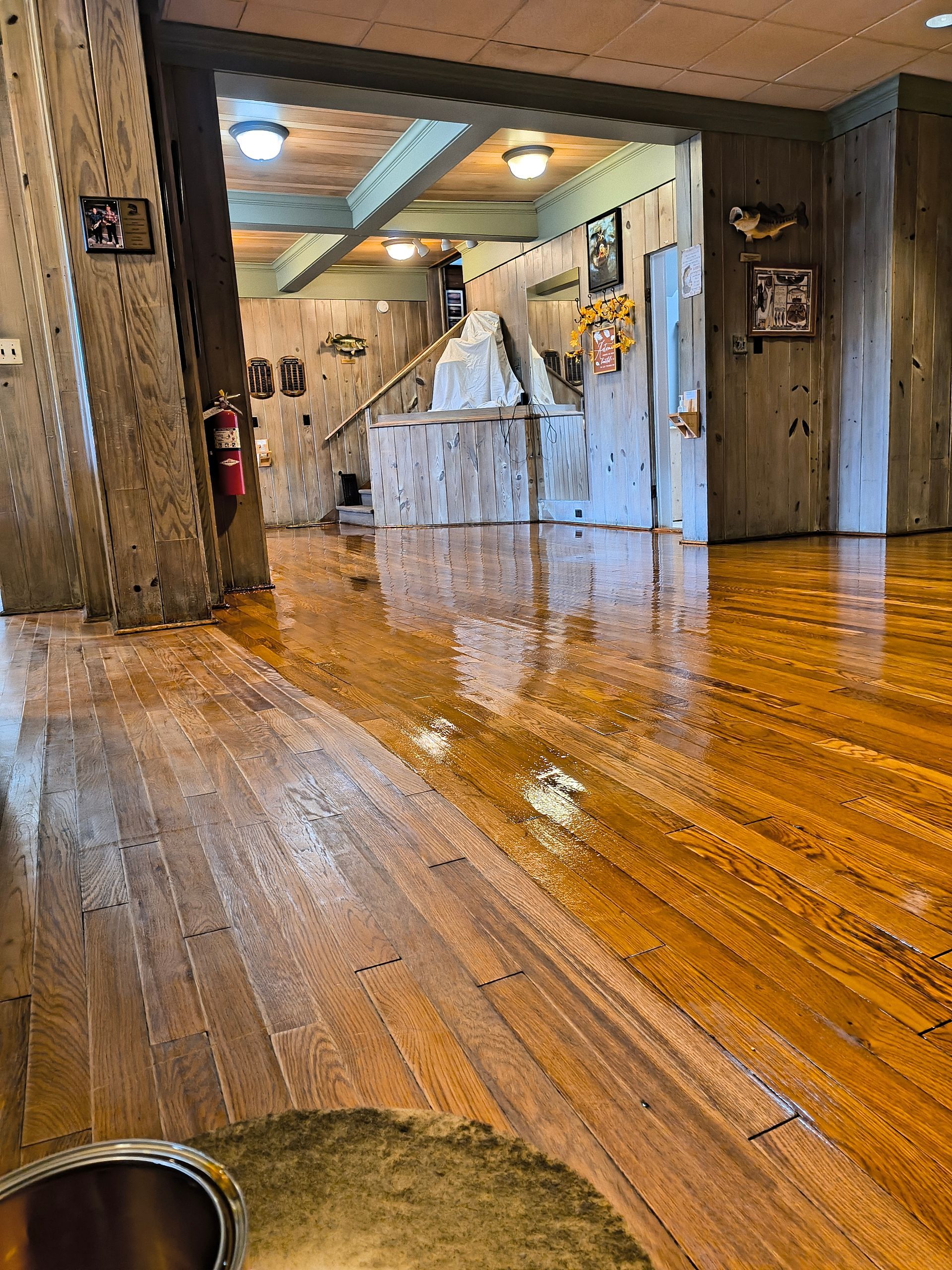 Wooden floor reflects water in a building with wood panel walls and a staircase visible in the distance.