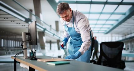 Man in blue apron sprays and cleans a desk in a modern office.
