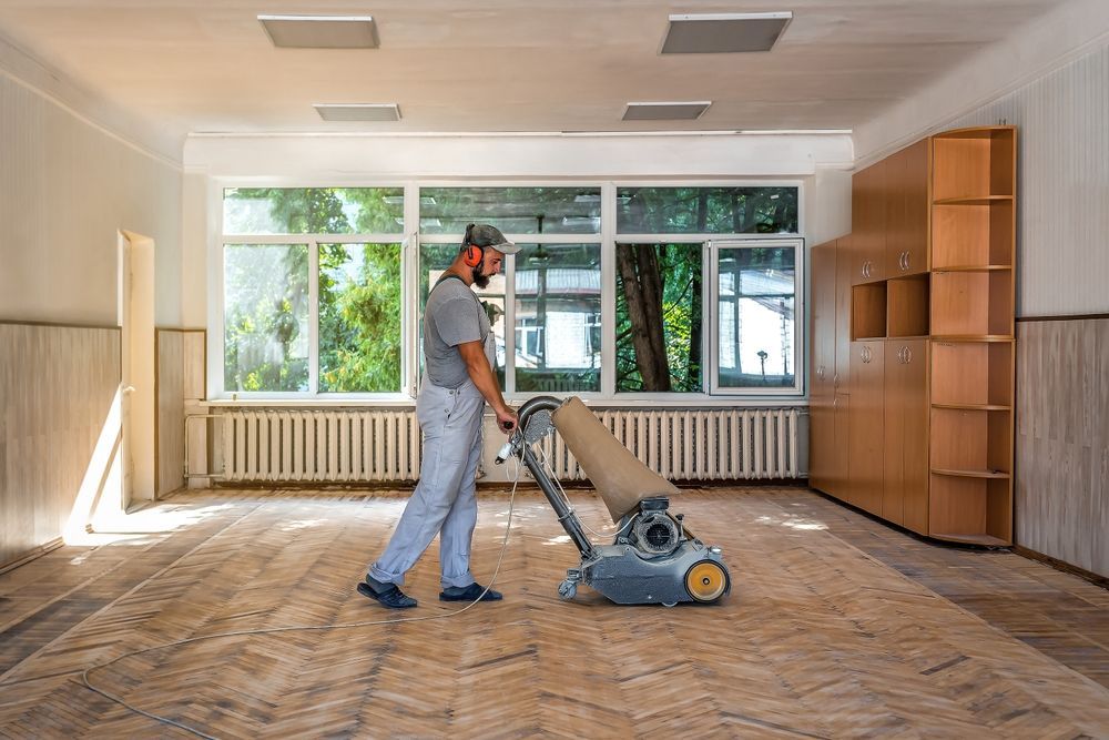 Person sanding a hardwood floor in a room with large windows, wearing a respirator.