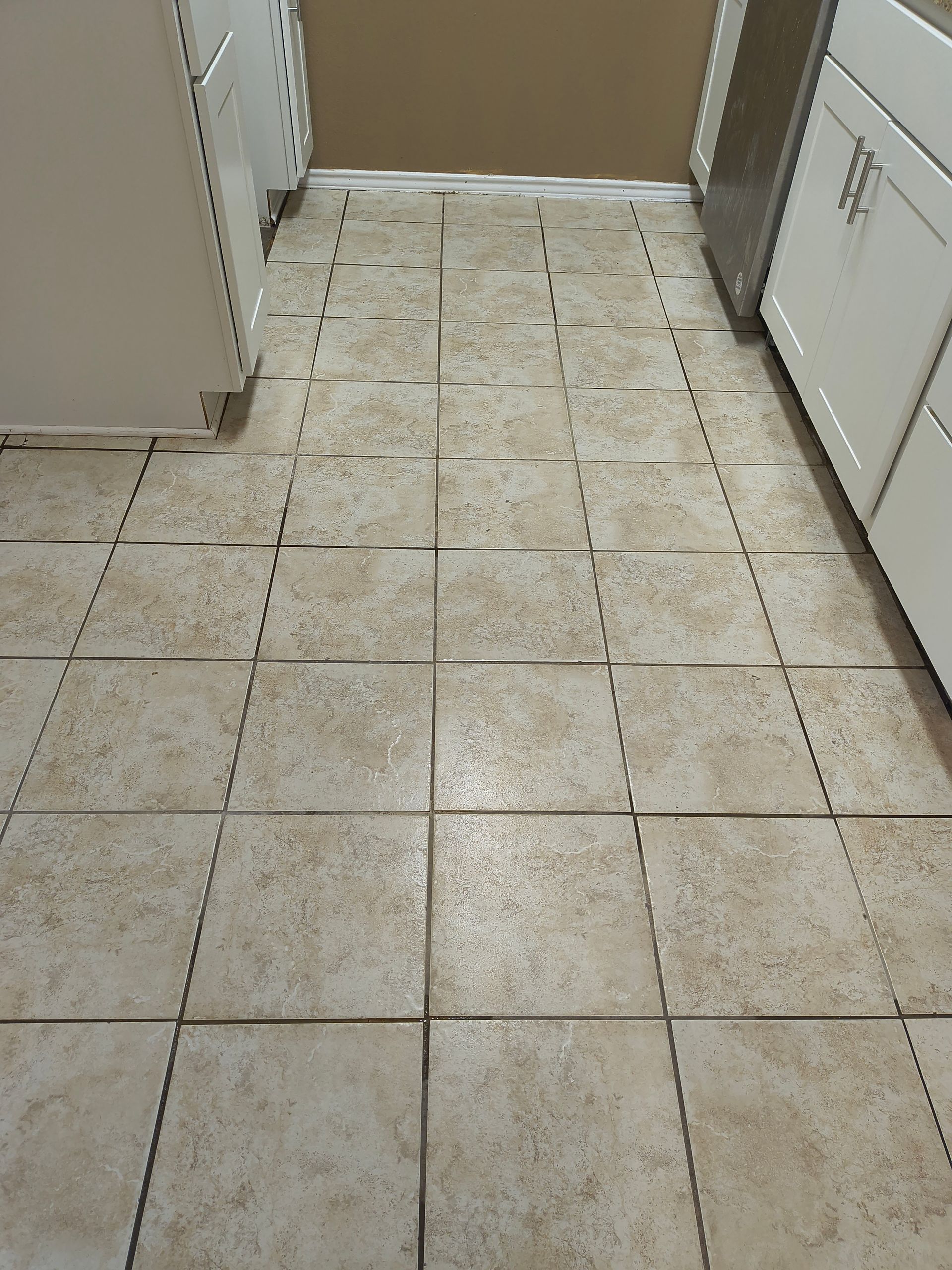 Tiled kitchen floor with several uneven tiles. Beige tiles with dark grout, between white cabinets and appliances.