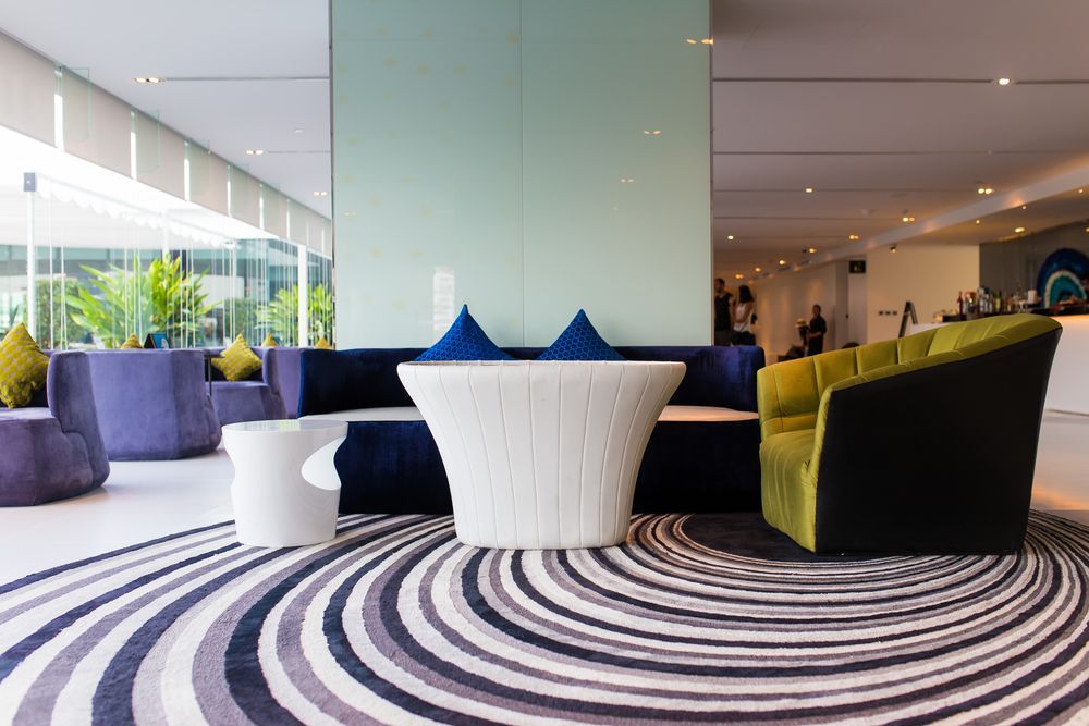 Modern hotel lobby with purple, blue, and green seating. Swirled patterned rug, bright lighting.